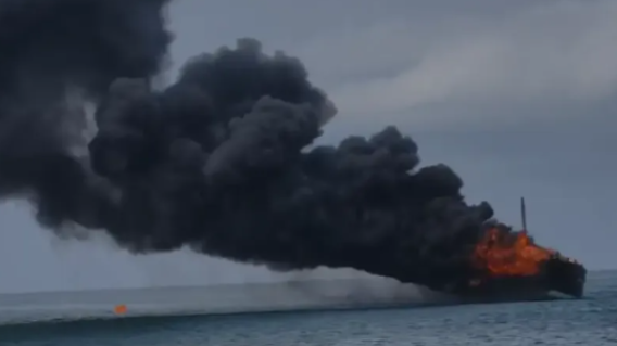 A cargo ship caught fire at the dock, with thick black smoke billowing into the sky.